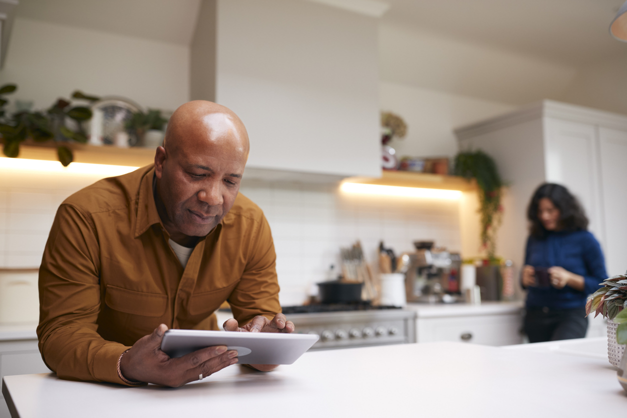 Mature Man At Home In Kitchen Looking At Digital Tablet With Female Partner In Background stock photo